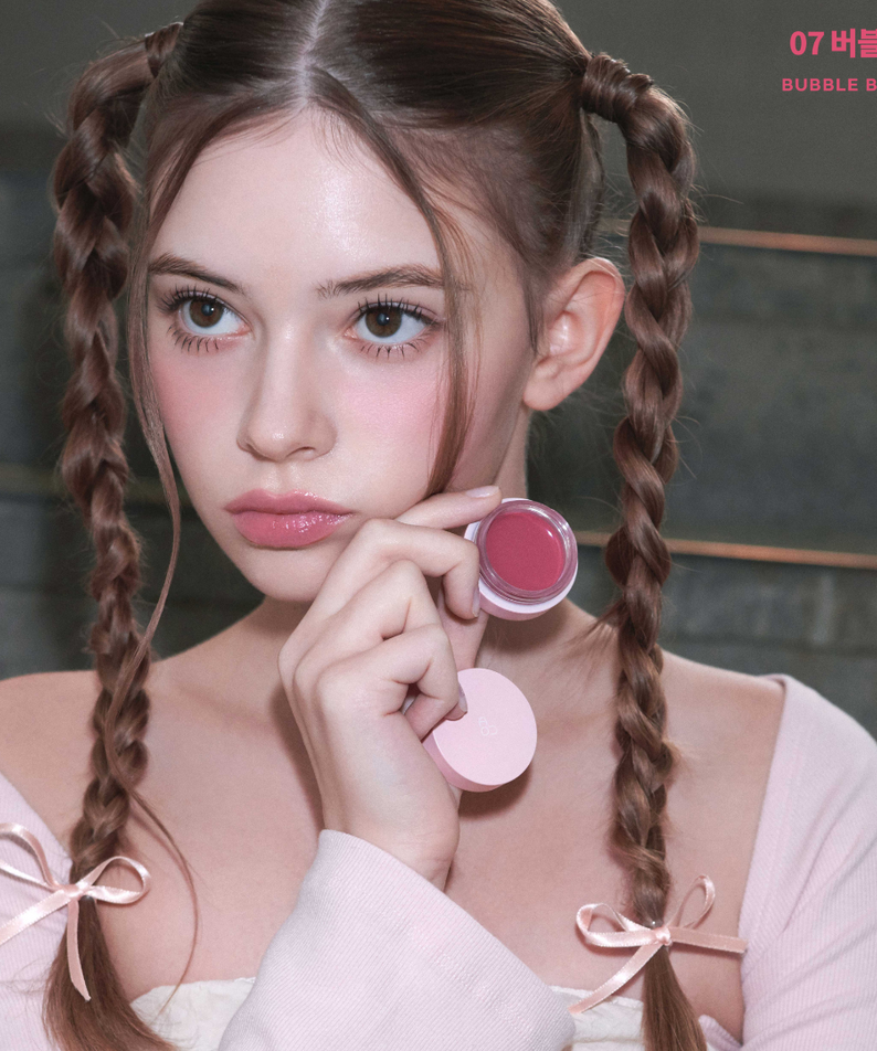 Jeune femme tenant le AOU Glowy Tint Balm, maquillage rosé pour lèvres, ambiance douce et élégante en arrière-plan.