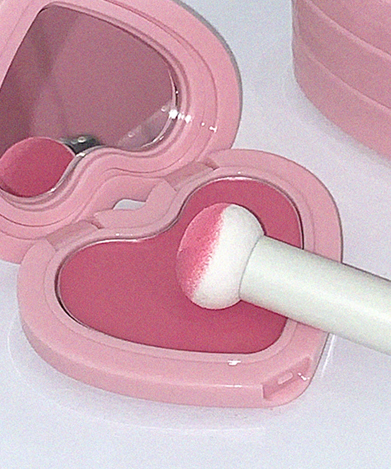 AOU Soft Cream Blush dans un boîtier rose en forme de cœur avec applicateur mousse. Maquillage crémeux pour un teint éclatant.
