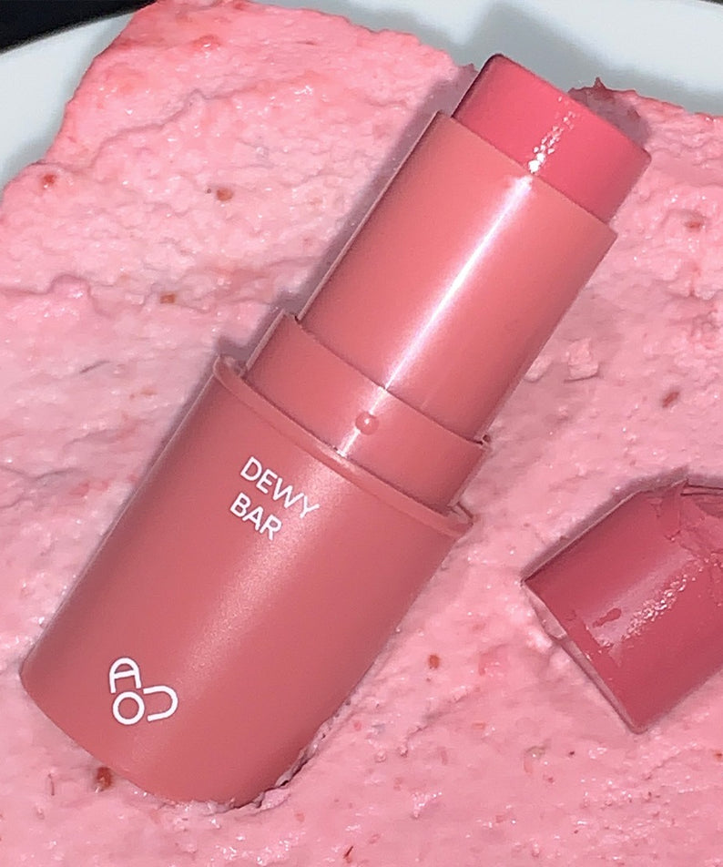 AOU Dewy Bar sur une texture rose crémeuse, représentant l'hydratation et la brillance naturelles du produit.