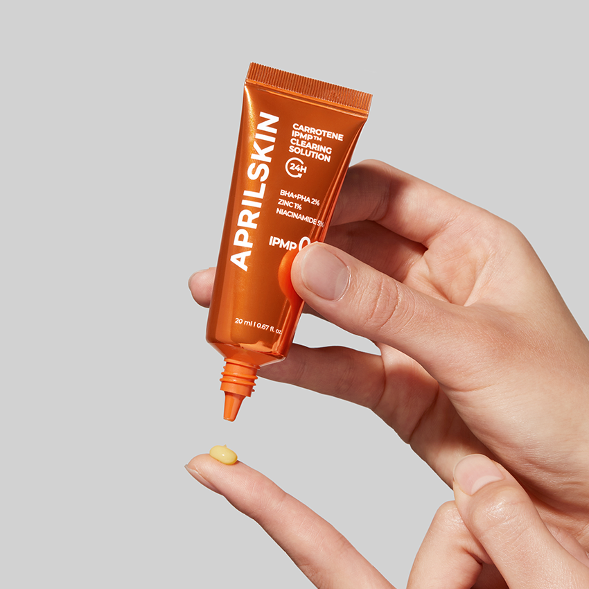 Décris l'image en français : APRILSKIN Carrotene IPMP Clearing Solution d’APRILSKIN, tube orange sur main tenant un échantillon jaune.