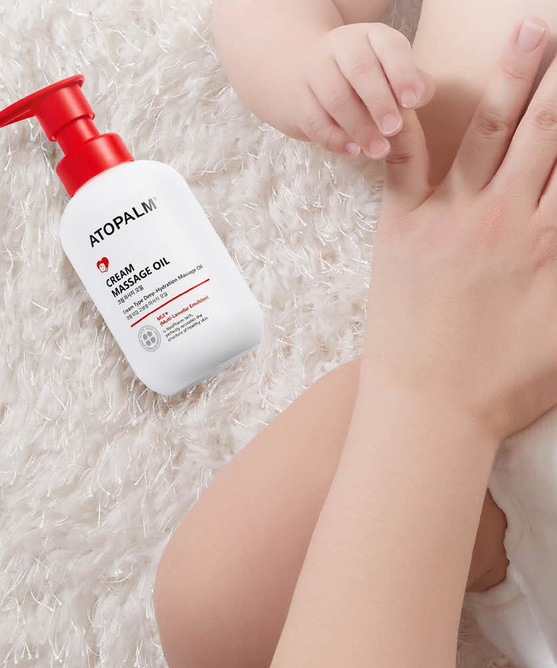 Une main massant bébé avec ATOPALM Cream Massage Oil 200ml, soin adapté pour la peau délicate, bouteille posée sur le lit moelleux.