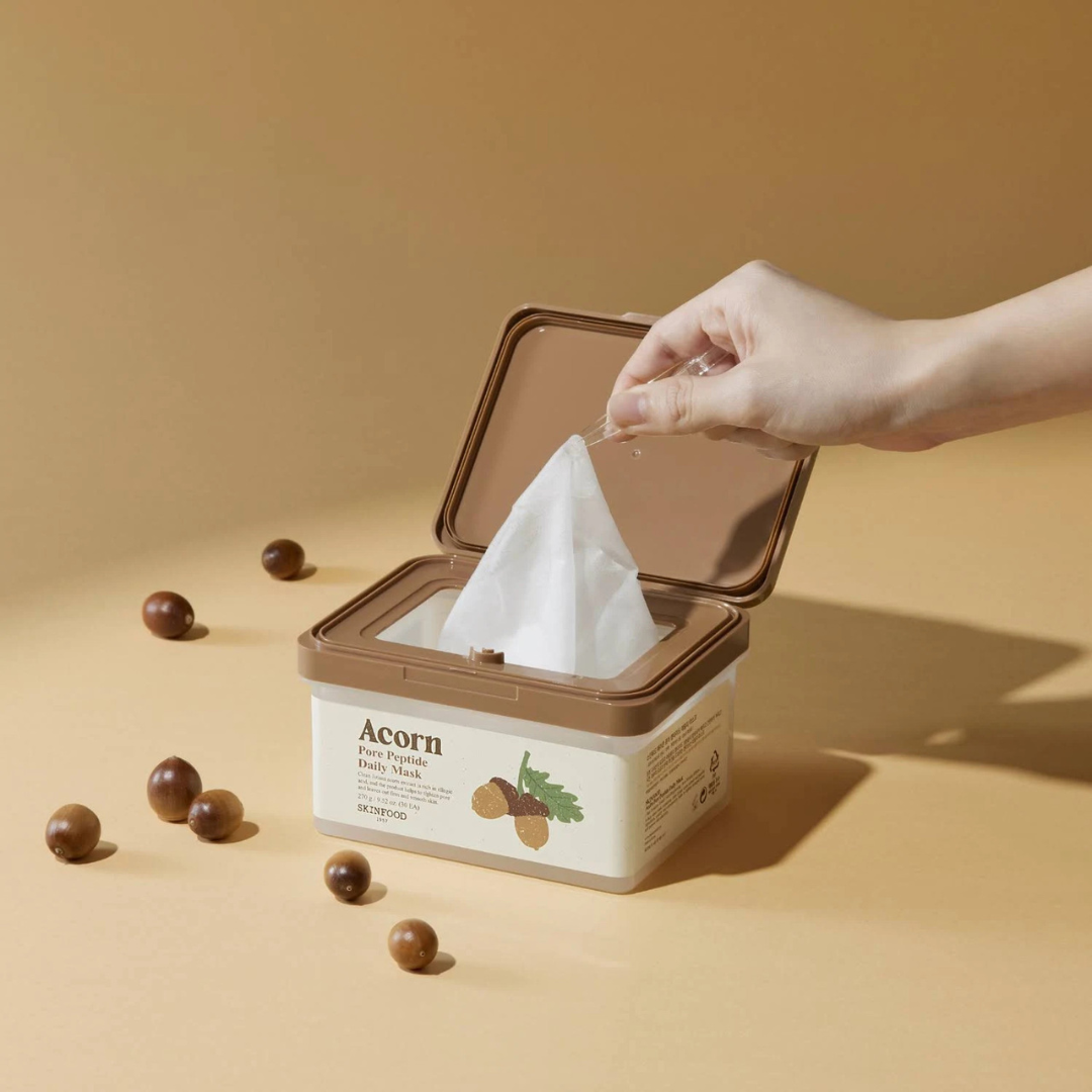 SKINFOOD Acorn Pore Peptide Daily Mask 30p 270g, boîte ouverte sur fond beige, main retire un tissu masque, contexte visuel spa.