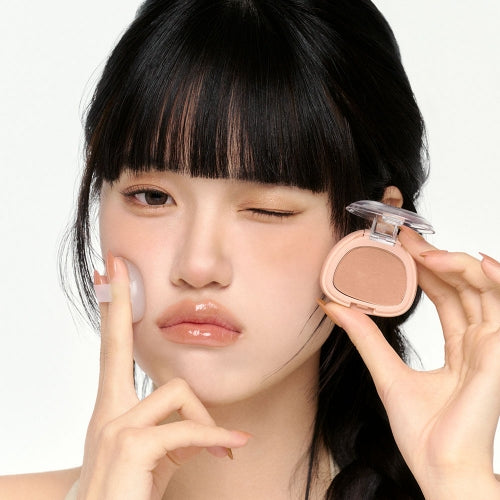 BBIA Perfect Match Blush de BBIA, maquillage appliqué sur le visage en gros plan, fond clair, mains tenant le blush