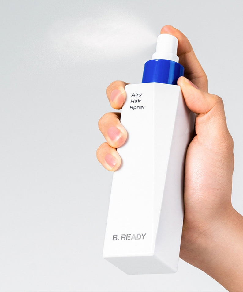 Main tenant un flacon de BE.READY Airy Hair Spray 200ml, vaporisateur à la main, sur fond clair.