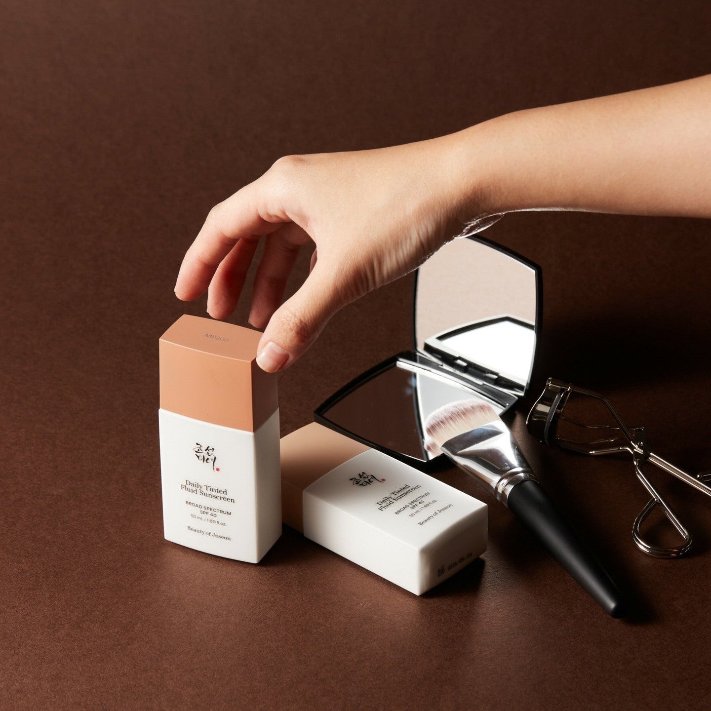 BEAUTY OF JOSEON Daily Tinted Fluid Sunscreen 50ml, pack sur fond brun, main appliquant un fluide teinté, marque visible.