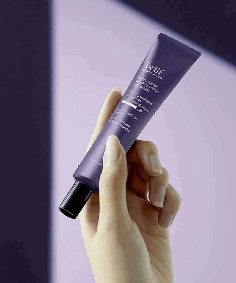 Tube de BELIF Age Knockdown V Eye Serum 25ml, tenue par une main, sur fond violet, produit de soin des yeux anti-âge.