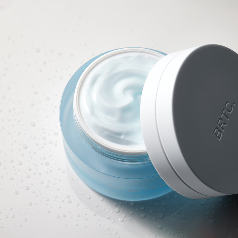BRTC Hydra Daily Cream 50ml de BRTC, crème hydratante bleue en pot ouvert sur fond blanc avec gouttes d’eau.