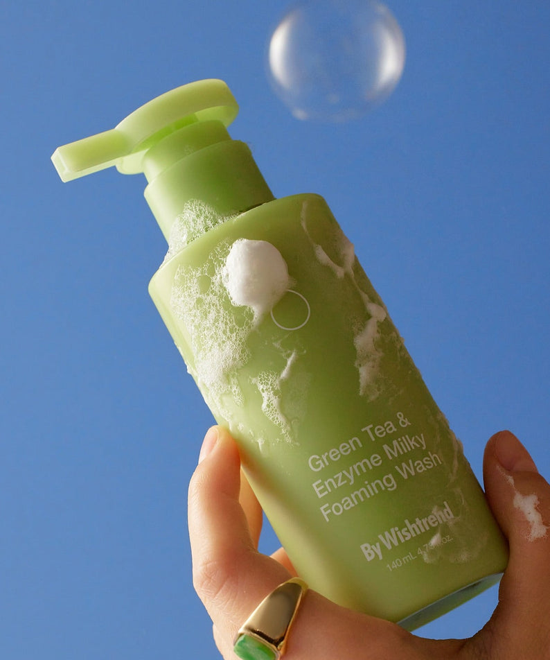 BY WISHTREND Green Tea & Enzyme Milky Foaming Wash 140ml : flacon vert en main, mousse sur fond bleu.