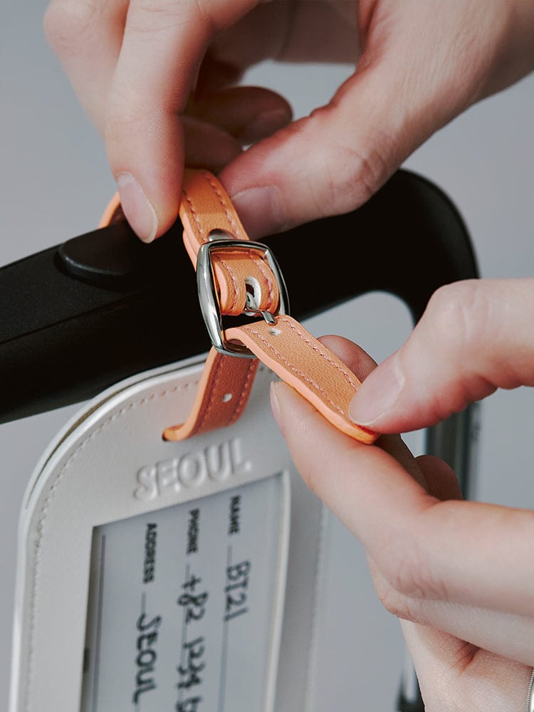 BT21 LUGGAGE TAG SEOUL EDITION : main accrochant l’étiquette de valise officielle LINE FRIENDS, fond valise “SEOUL”
