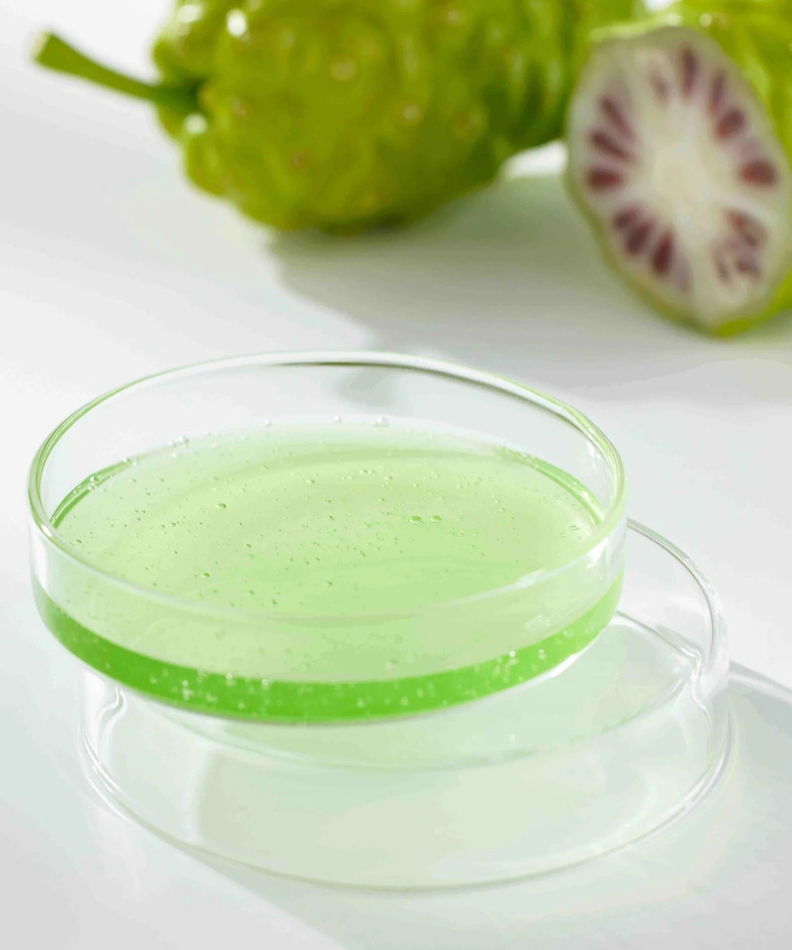 CELIMAX Noni Ampoule Calming+Radiance 100ml dans une coupelle en verre avec un fruit de noni en arrière-plan.