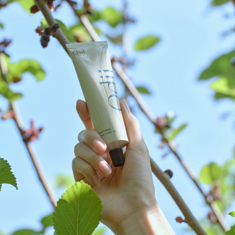 Main tenant CHWI (CHI) Odii Hand Cream 50ml, tube vert, feuilles et ciel bleu en arrière-plan, contexte naturel.