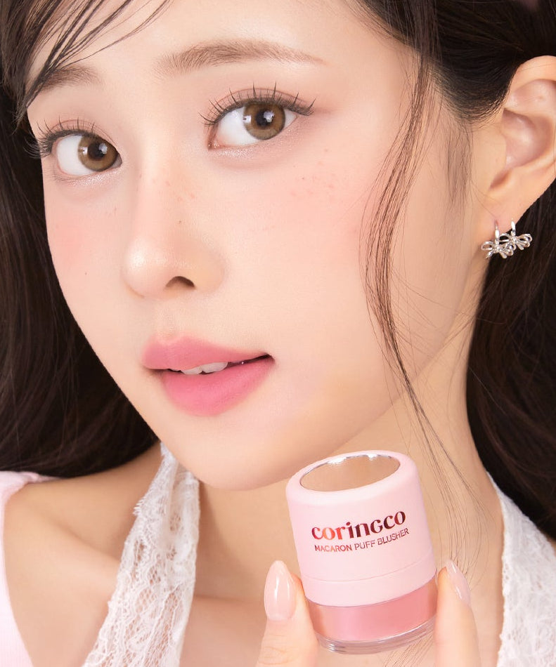 Décris l'image en français : CORINGCO Macaron Puff Blusher, marque Coringco, sur visage maquillé, main tenant le produit.