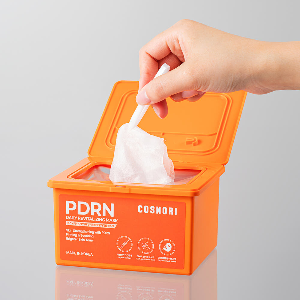 Main tenant COSNORI PDRN Daily Revitalizing Mask 30 sheets, boîte orange avec les lingettes masques PDRN, contexte cosmétique