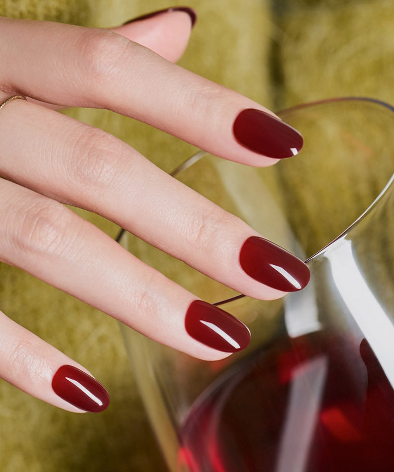 DASHING DIVA  Classic Burgundy (Oval) : ongles bordeaux ovales, main près d’un verre, contexte visuel en extérieur, marque DASHING DIVA.