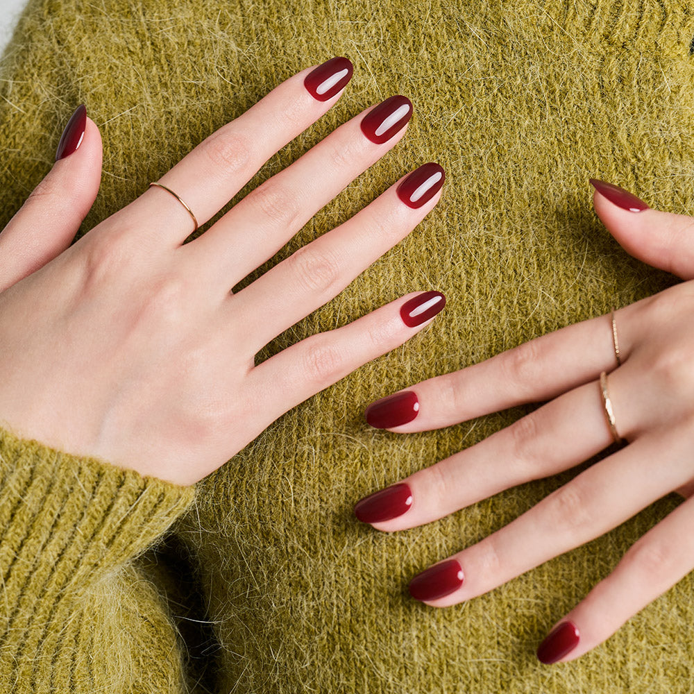 DASHING DIVA  Classic Burgundy (Oval), manucure ovale sur mains posées sur un tissu vert, ambiance mode.