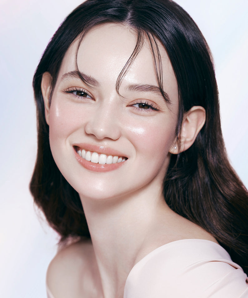 Femme souriante portant DEAR DAHLIA Prime Layer Glow Primer avec finition lumineuse et radieuse, produit de beauté haute qualité.