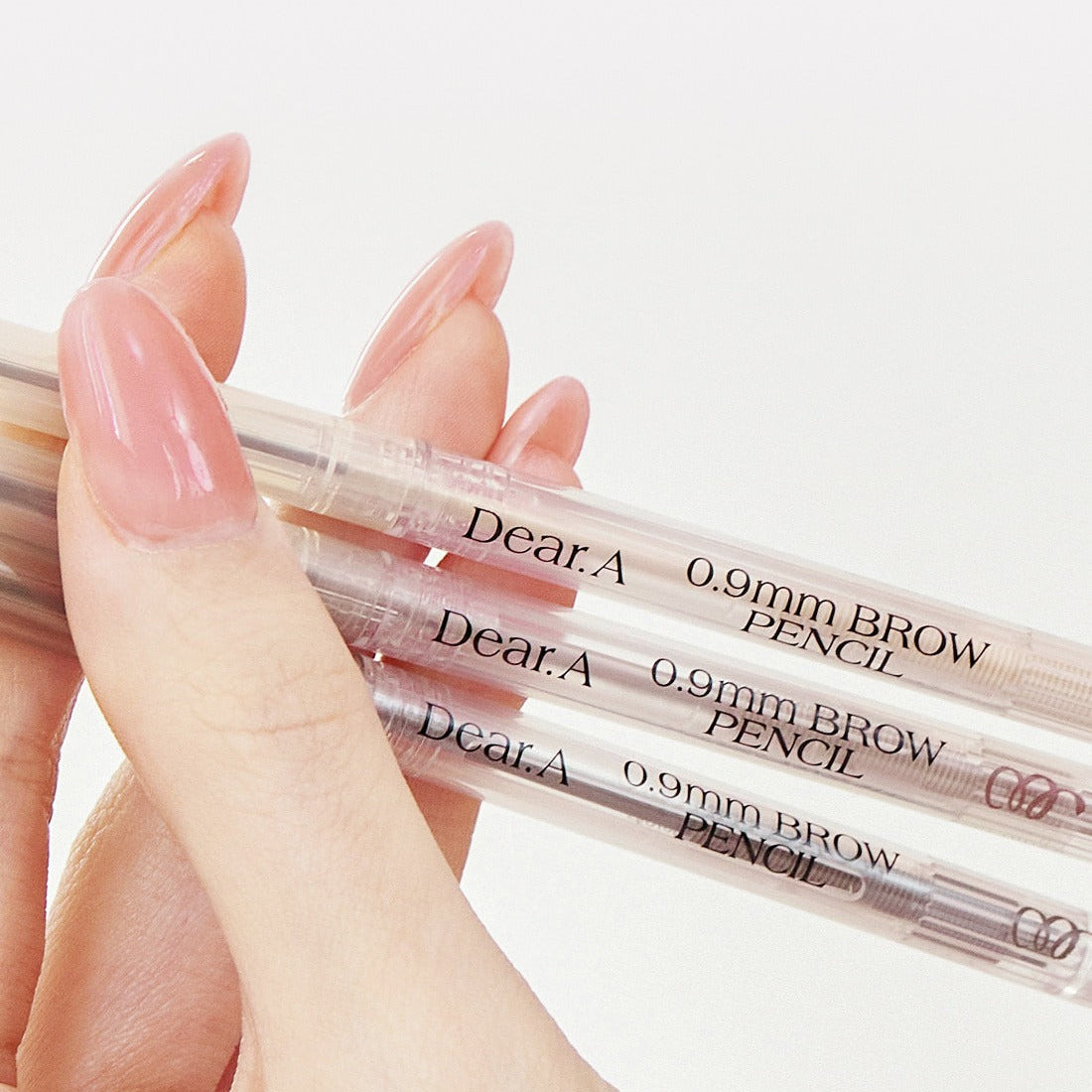 Main tenant DEAR.A 0.9mm Eyebrow, stylo anti-cernes, marques lisibles sur produit transparent, fond clair.