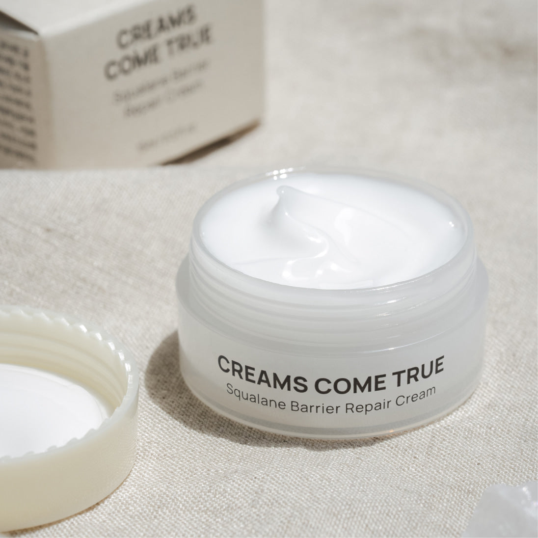 DE:MAF Creamscumtrue Squalane Barrier Repair Cream 15ml, crème blanche dans pot, sur tissu beige, marque visible.