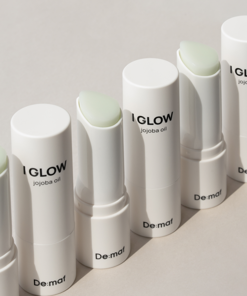 Décris l'image en français en mentionnant le produit, la marque et le contexte visuel: DE:MAF Eye Glow Jojoba Oil Multi Balm 9g, baumes tubes blancs sur fond clair.