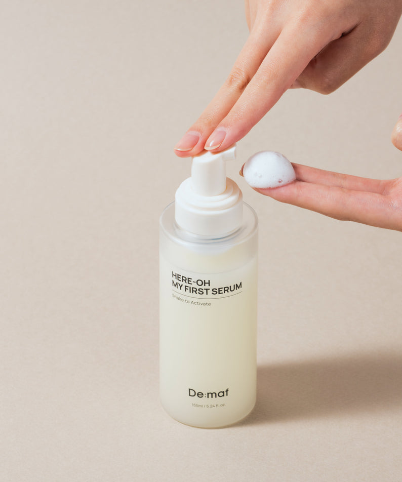 DE:MAF Here-Oh My First Serum 155ml, main ouvrant une pompe, produit De:maf sur fond beige, contexte soin beauté.