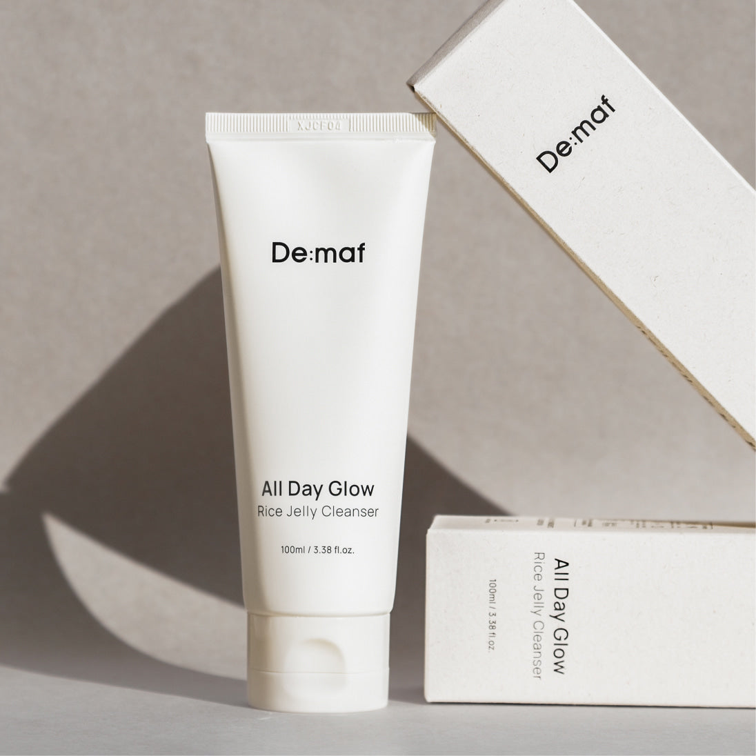 DE:MAF Rice Jelly Cleanser 100ml De:maf, tube blanc sur fond beige avec coffret en arrière-plan.