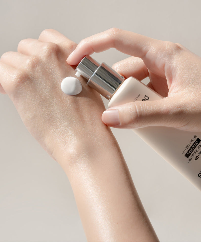 Décris l'image en français : DE:MAF Sun Gloss Glow Up Inorganic Sunscreen 50ml Original, tube sur une main, texture crème.