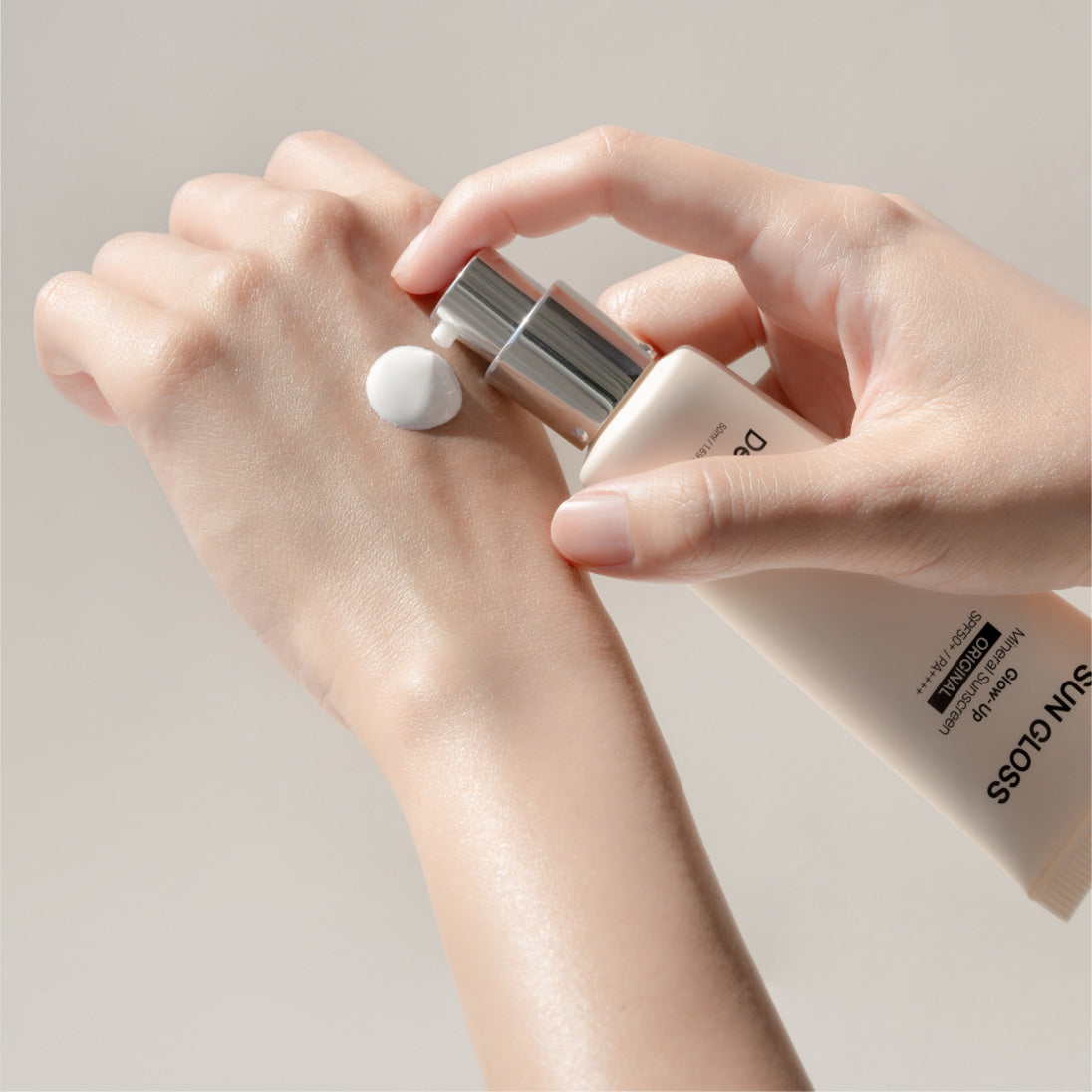 Décris l'image en français : DE:MAF Sun Gloss Glow Up Inorganic Sunscreen 50ml Original, tube sur une main, texture crème.