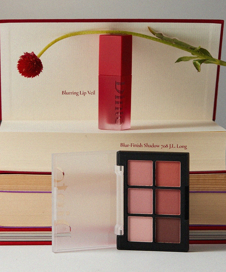 Emballage DINTO Blurring Lip Veil avec palette de fards, posé sur des livres artistiquement, maquillage élégant et moderne.