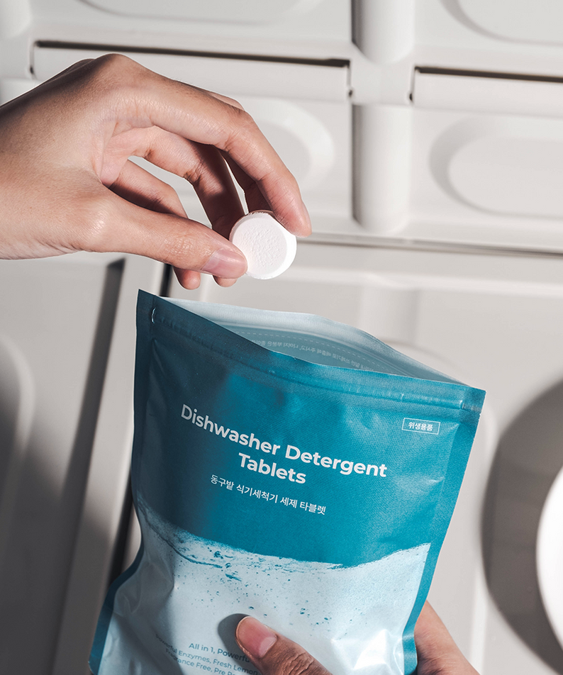 Main versant un comprimé dans le sac DONGGUBAT Dishwasher Detergent Tablets 60 240g, sur fond de cuisine moderne.