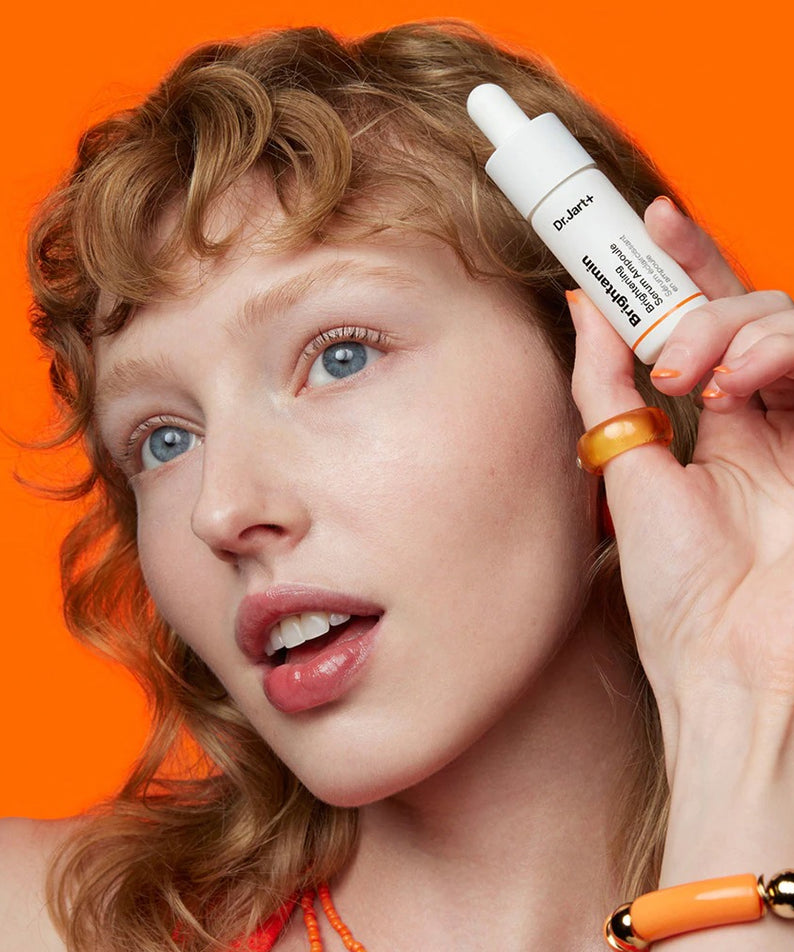 Femme posant avec le sérum DR.JART+ Brightamine Brightening Serum Ampoule sur un fond orange vibrant.