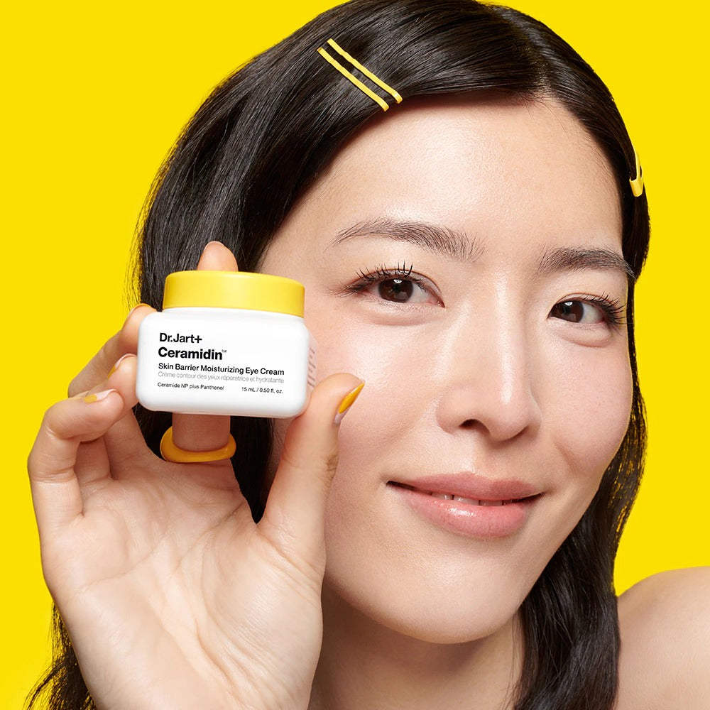 Décris l'image en français : DR.JART+ Ceramidin Skin Barrier Moisturizing Eye Cream 15ml, femme montrant le pot sur fond jaune.