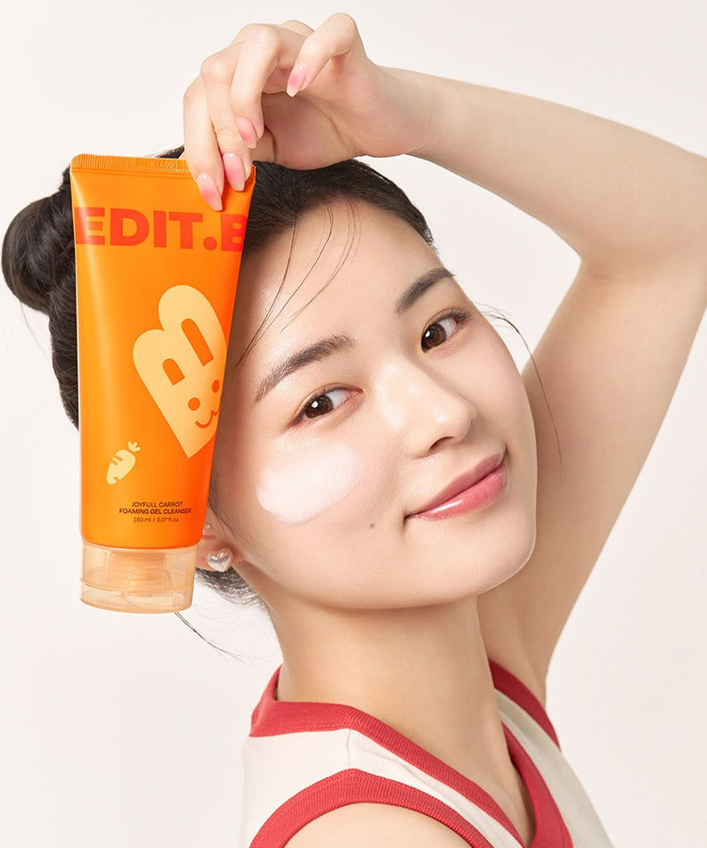 Jeune femme souriante tenant un tube de 150ml d'EDIT.B Joyful Carrot Foaming Gel Cleanser, soin nettoyant visage.