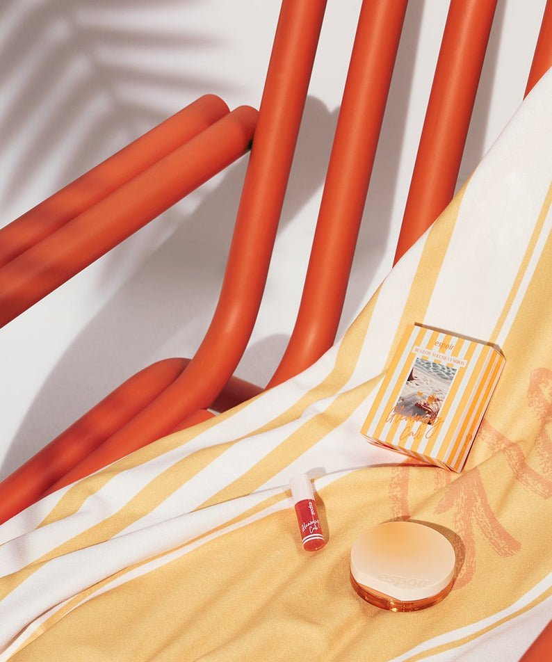 Coussin ESPOIR BE GLOW VOLUME CUSHION SPF42 PA+++ [Gleaming CALI Edition] sur chaise orange, avec accessoires beauté.