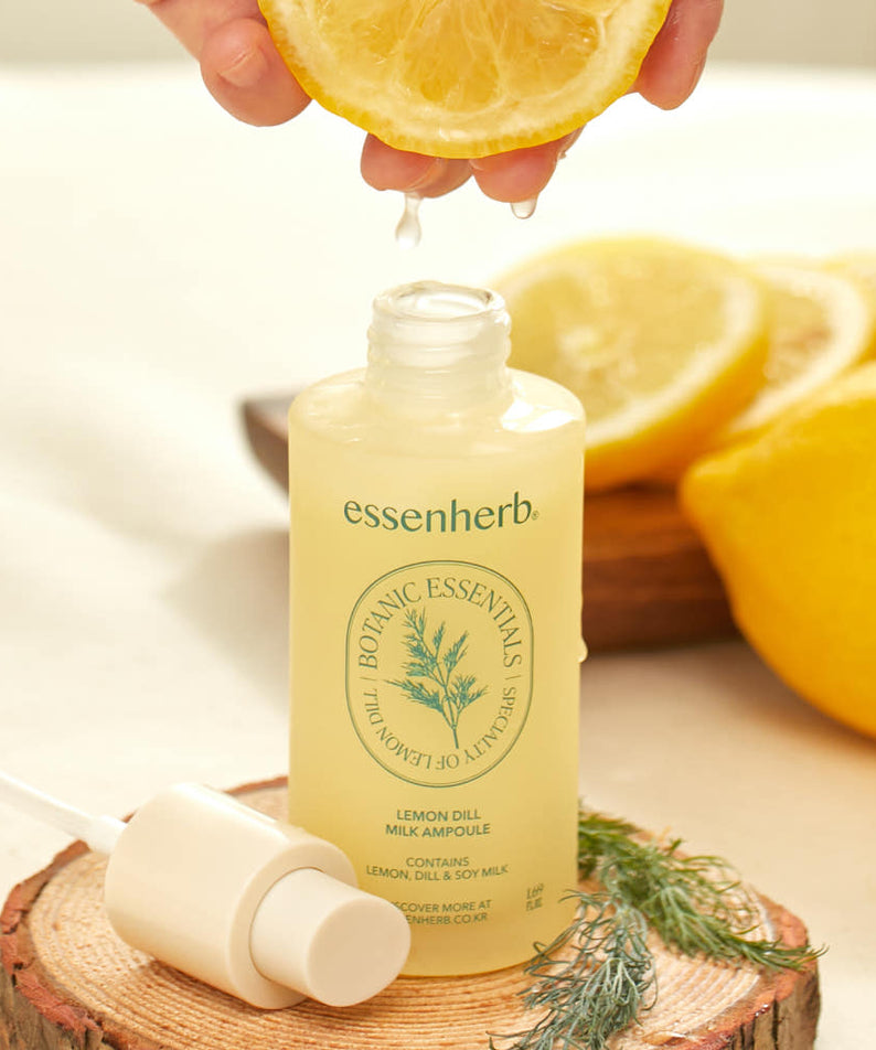 ESSENHERB Lemon Dill Milk Ampoule 50ml, flacon posé sur bois, avec citron et brin d’aneth, main vers une goutte.