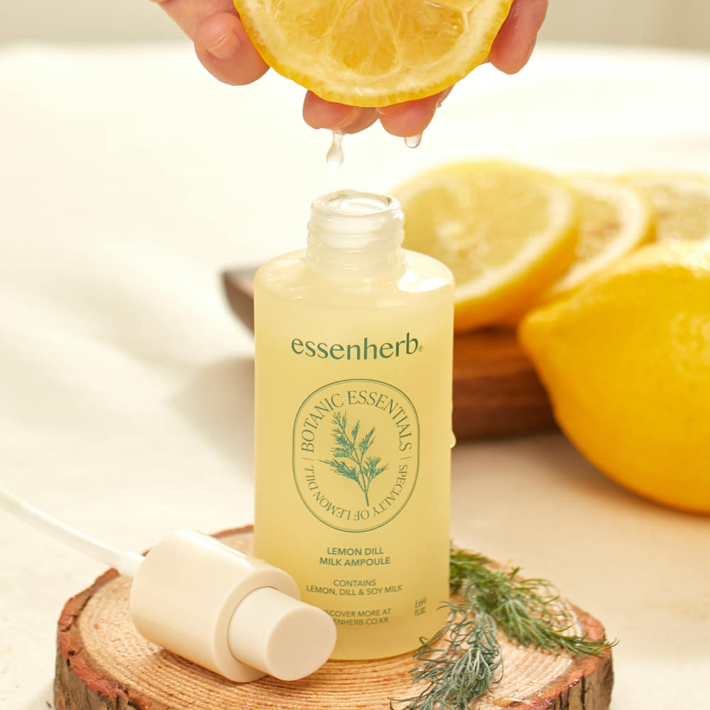 ESSENHERB Lemon Dill Milk Ampoule 50ml, flacon posé sur bois, avec citron et brin d’aneth, main vers une goutte.