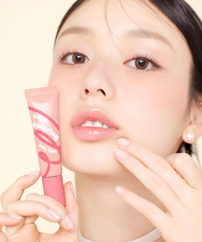 Décris l'image en français : ETUDE  Sugar Coloring Jelly Balm 9g, tube maquillage; femme appliquant un baume colorant, fond clair.