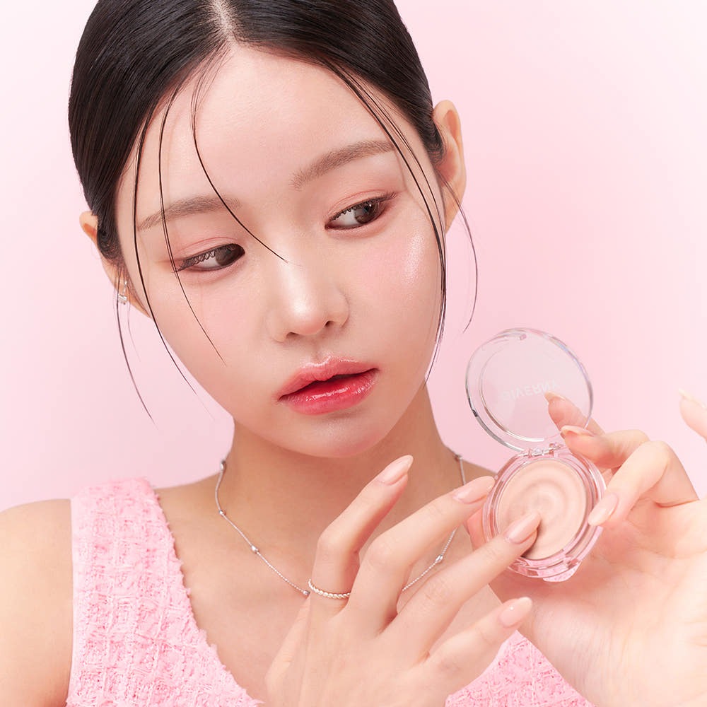 GIVERNY Dewy Voluming Highlighter 3g de la marque GIVERNY, femme applique un enlumineur lumineux en gros plan, fond rose.
