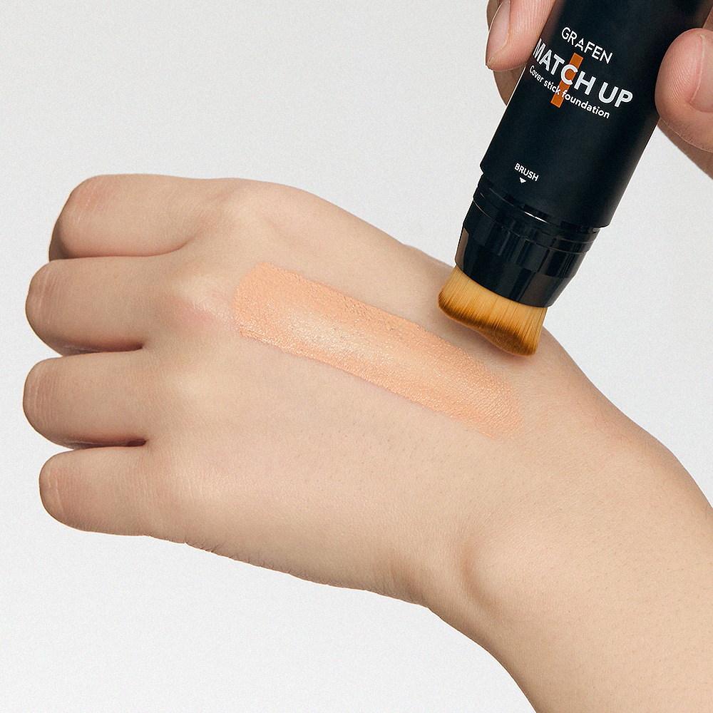 GRAFEN  Match Up Cover Stick Foundation 12g : main appliquant un fond de teint stick avec swatch sur peau, fond clair.