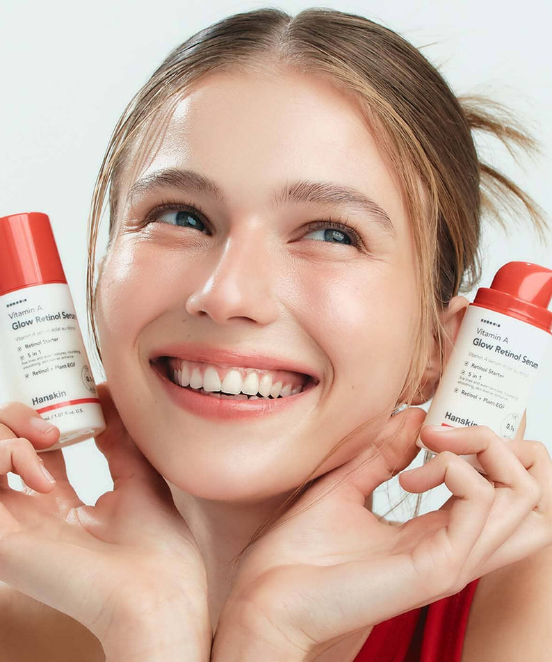 Décris l'image en français : HANSKIN Vitamin A Glow Retinol Serum 30ml, marque HANSKIN, femme souriante tient deux flacons.