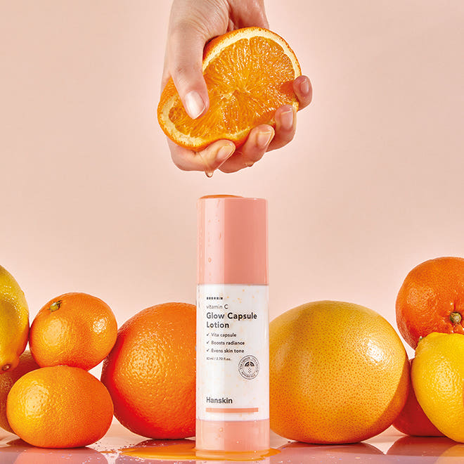 HANSKIN Vitamin C Glow Capsule Lotion 80ml : flacon rose entre oranges, main tenant une orange, éclairage studio sur fond clair.