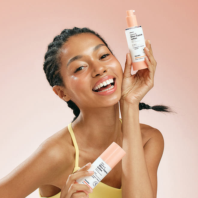 HANSKIN Vitamin C Glow Capsule Lotion 80ml, sur fond rose : femme souriante tient la lotion, marque HANSKIN en évidence.