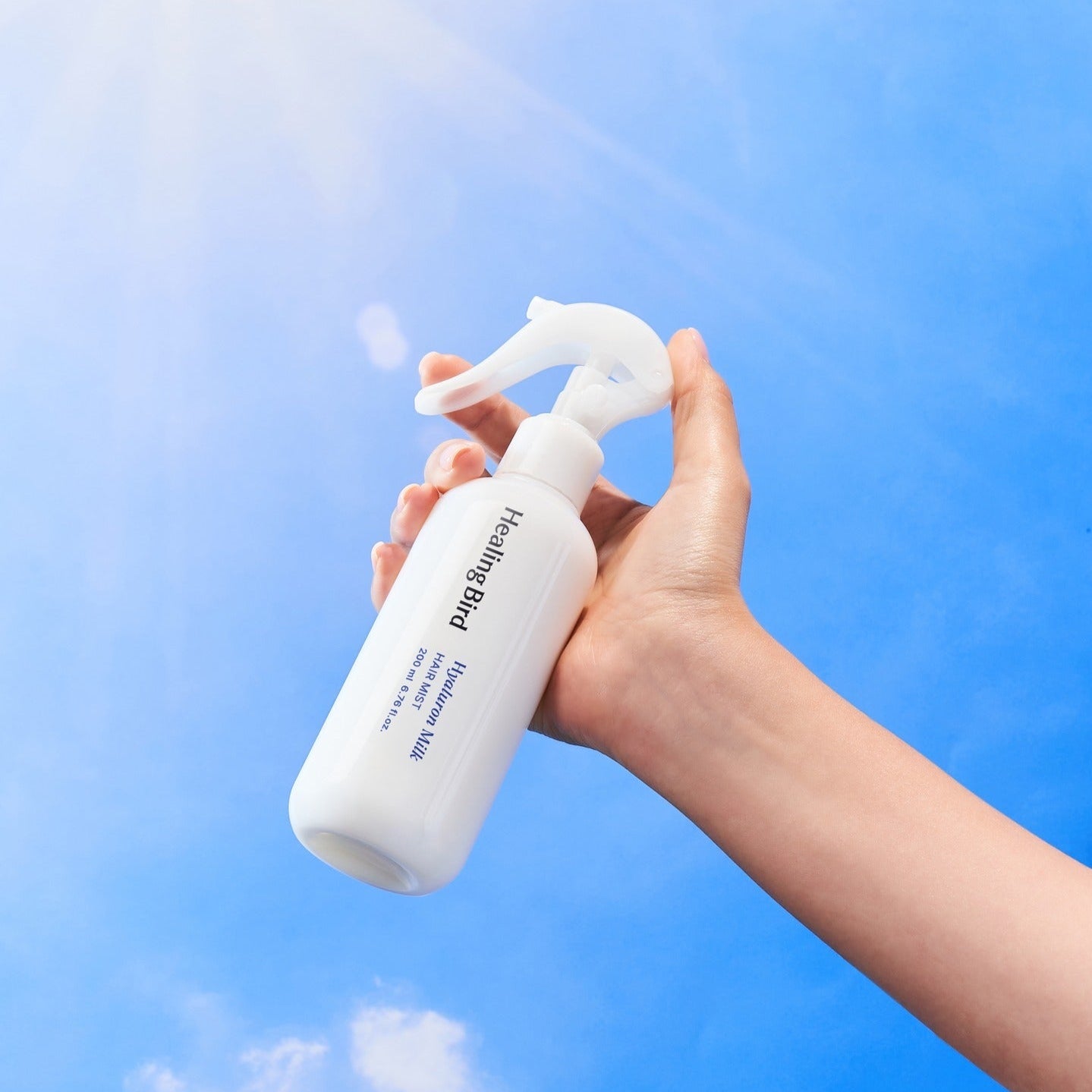 Main tenant HEALING BIRD Hyalutonic Milk Hair Mist 200ml, marque sur flacon, fond ciel bleu, spray cheveux, cosmétique