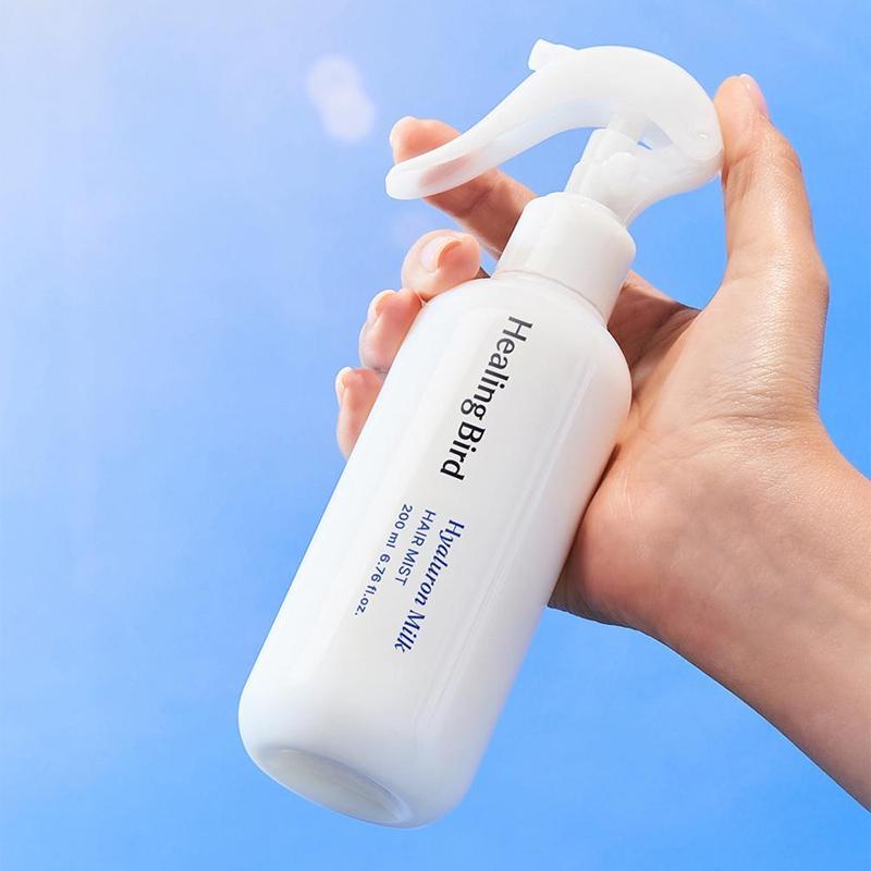 Main tenant HEALING BIRD Hyalutonic Milk Hair Mist 200ml, bouteille blanche avec spray, fond bleu.