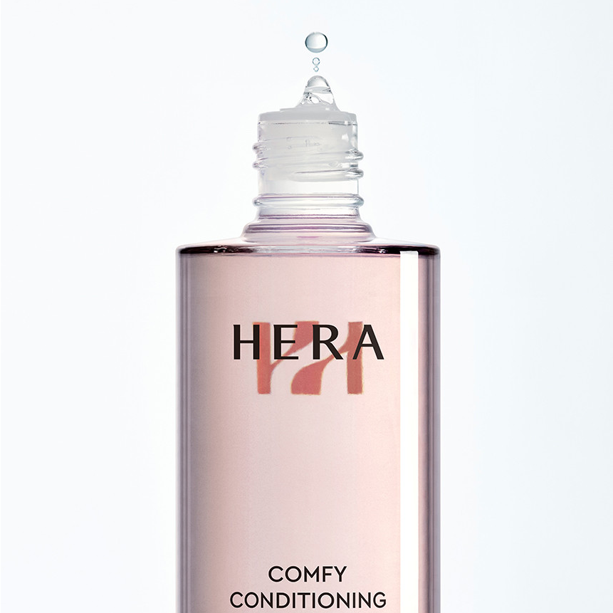 HERA  Comfy Conditioning Essence 150g, flacon cosmétique rose sur fond blanc, marque HERA en gros plan.
