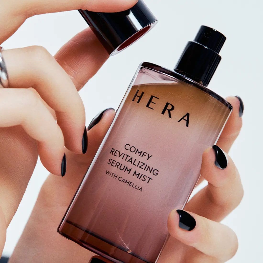 Décris l’image en français : HERA Comfy Revitalizing Serum Mist 90ml de la marque HERA, mains appliquant la brume sur flacon.