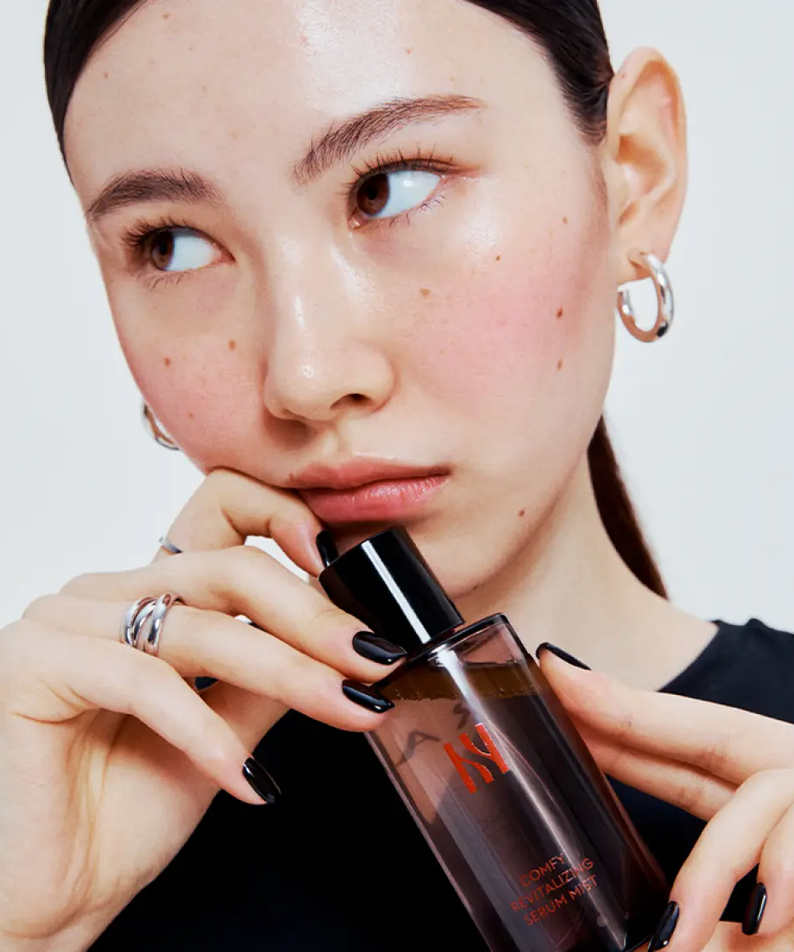 Décris l'image en français : HERA Comfy Revitalizing Serum Mist 90ml, femme l’utilisant sur peau, flacon HERA sur fond clair.
