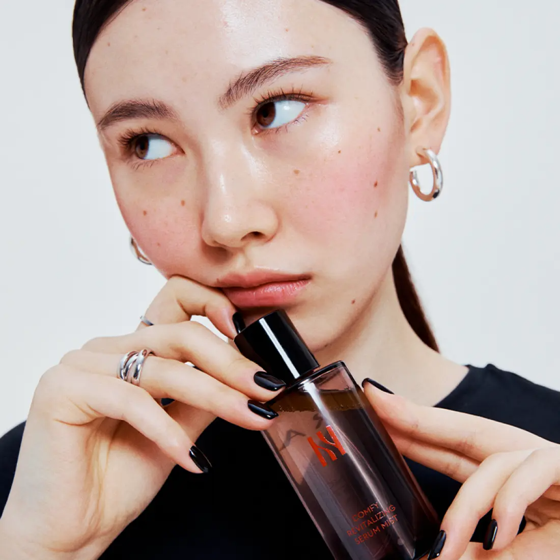 Décris l'image en français : HERA Comfy Revitalizing Serum Mist 90ml, femme l’utilisant sur peau, flacon HERA sur fond clair.