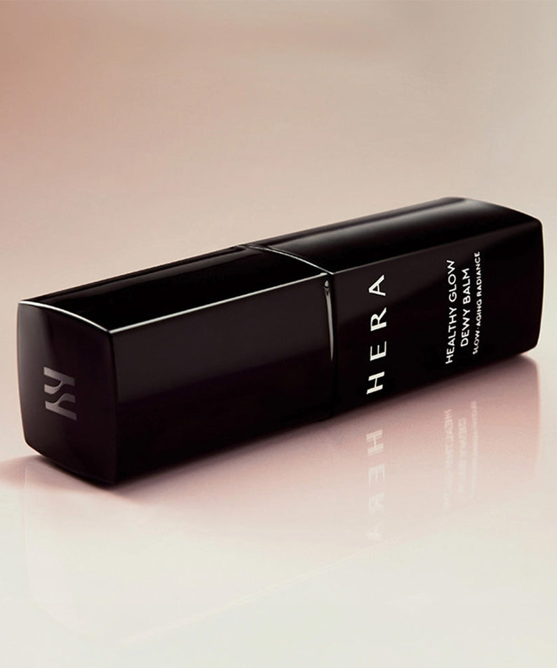 HERA  Healthy Glow Dewy Balm 10g, baume déwy HERA sur fond clair, packaging noir et reflet sur table