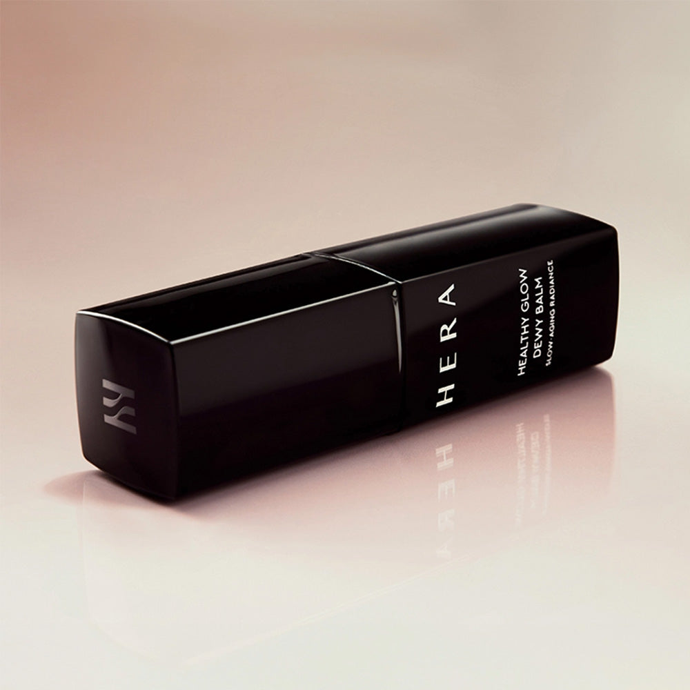 HERA  Healthy Glow Dewy Balm 10g, baume déwy HERA sur fond clair, packaging noir et reflet sur table