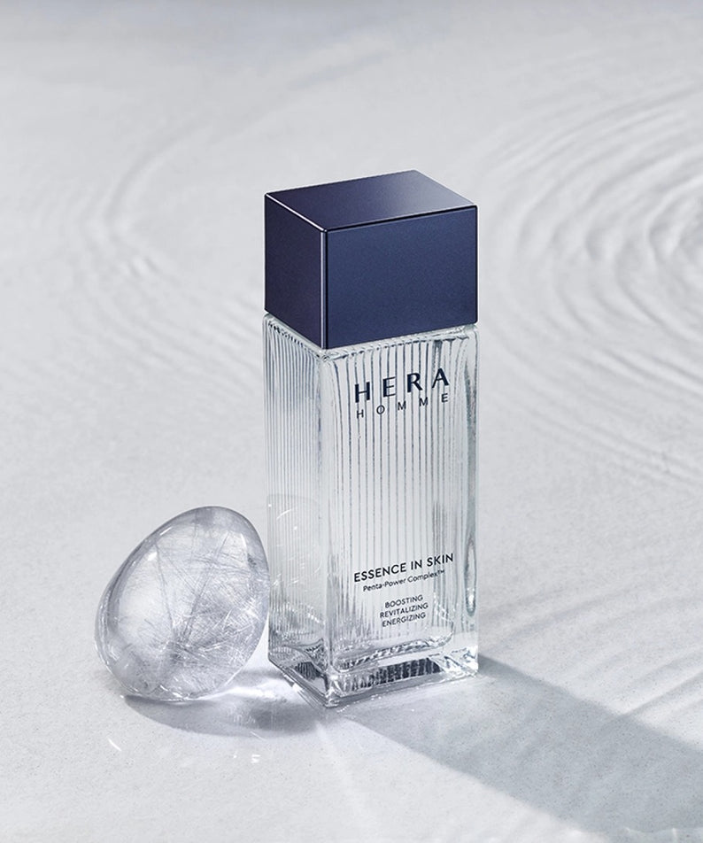 Décris l'image en mentionnant HERA Homme Essence In Skin 125ml, flacon HERA bleu sur fond blanc texture peau, contexte soin.